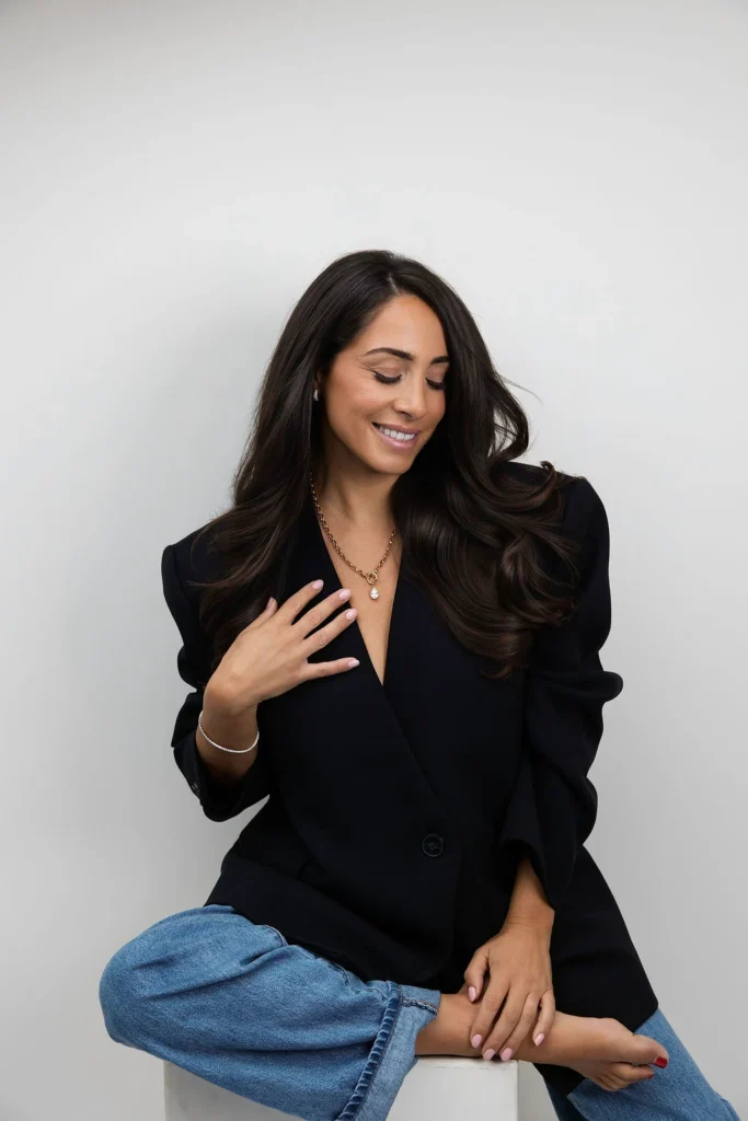 Woman with long dark hair wearing a black blazer and blue jeans sitting on a stool smiling softly with her hand resting on her chest