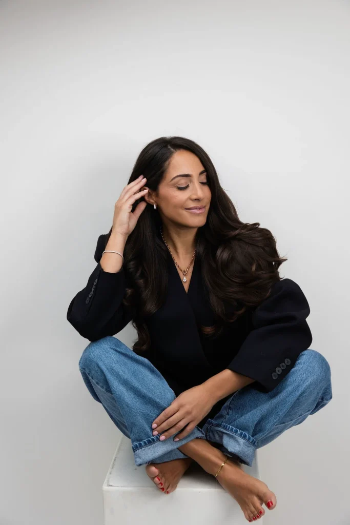 Woman sitting cross legged on a white pedestal against a light gray backdrop wearing a black blazer and blue jeans eyes closed in a calm pose