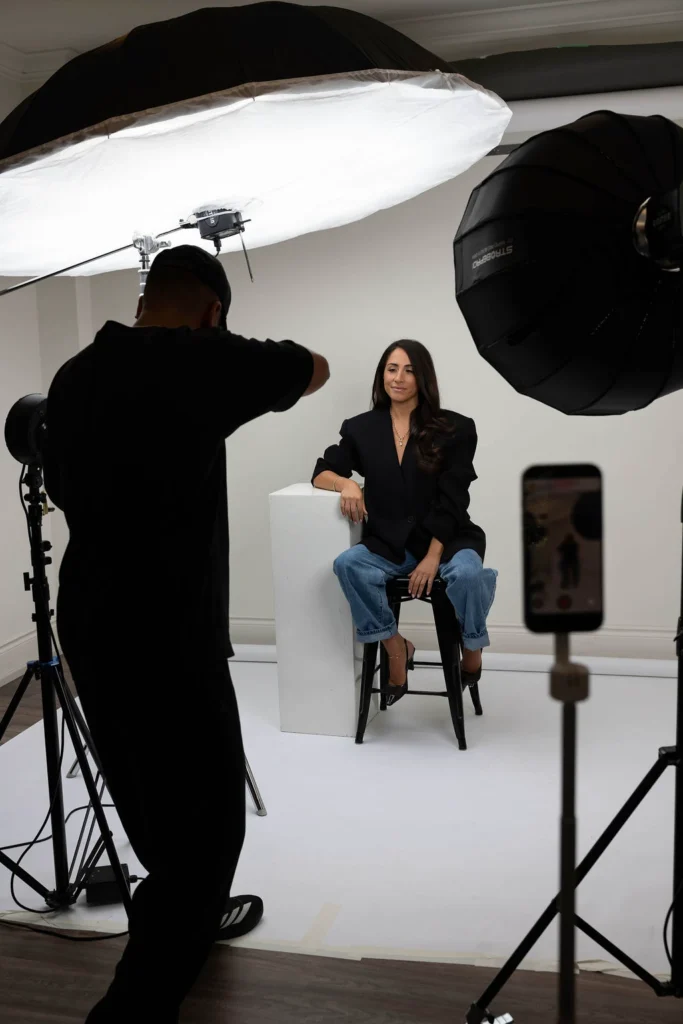 Studio photoshoot woman in a black blazer and jeans sits on a white block under large photography lights while a photographer adjusts equipment nearby
