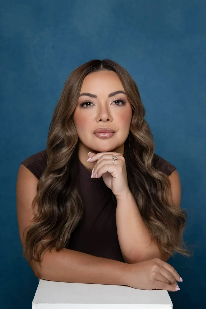 Portrait of a woman with long wavy brown hair resting her chin on her hand wearing a dark sleeveless top against a blue backdrop