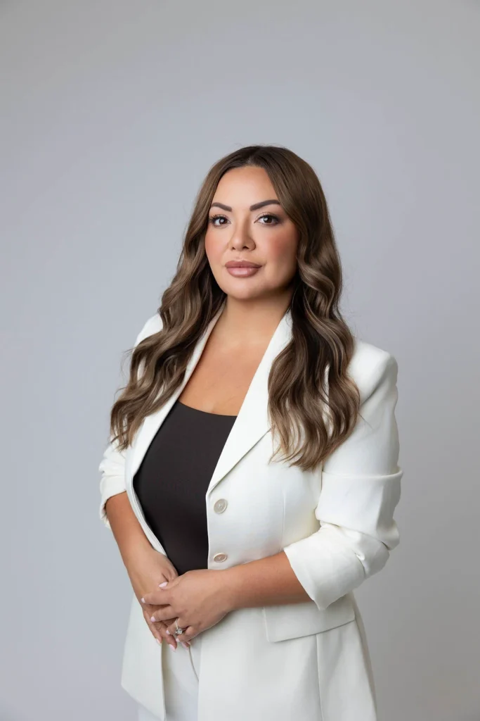 Portrait of a professional woman with long wavy brown hair wearing a white blazer over a black top against a gray studio backdrop