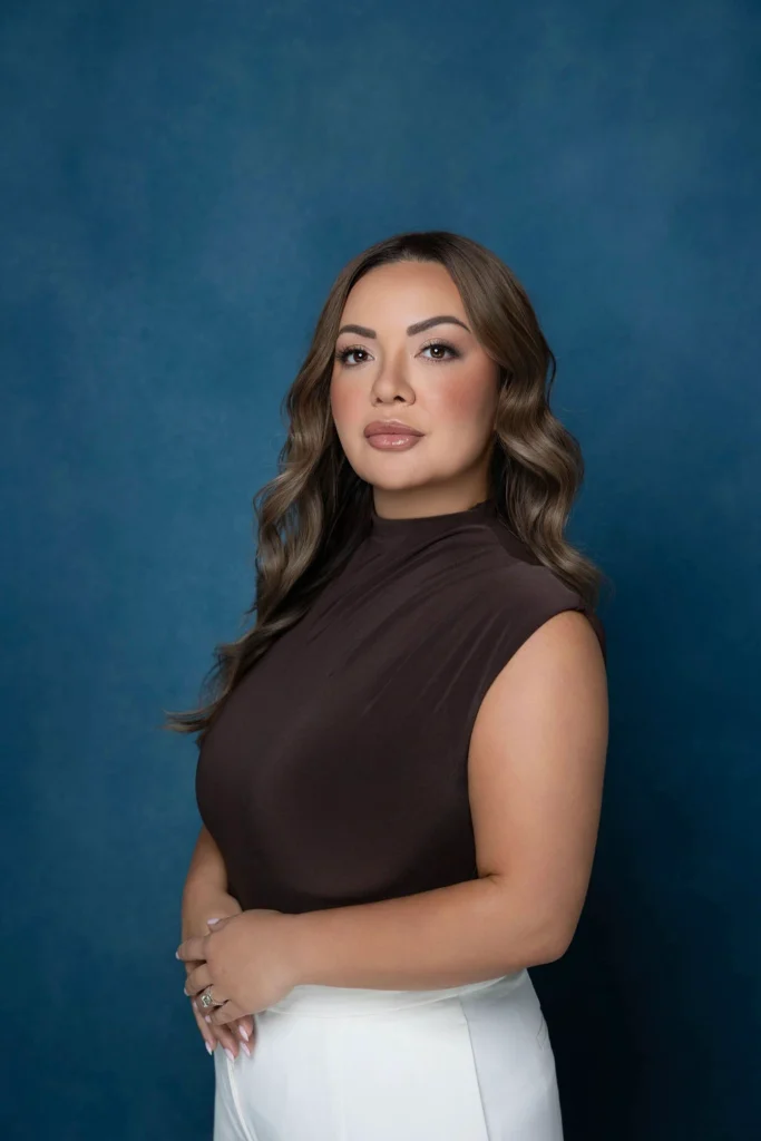 Professional headshot of a woman with wavy hair wearing a sleeveless brown top and white pants posed against a blue studio backdrop