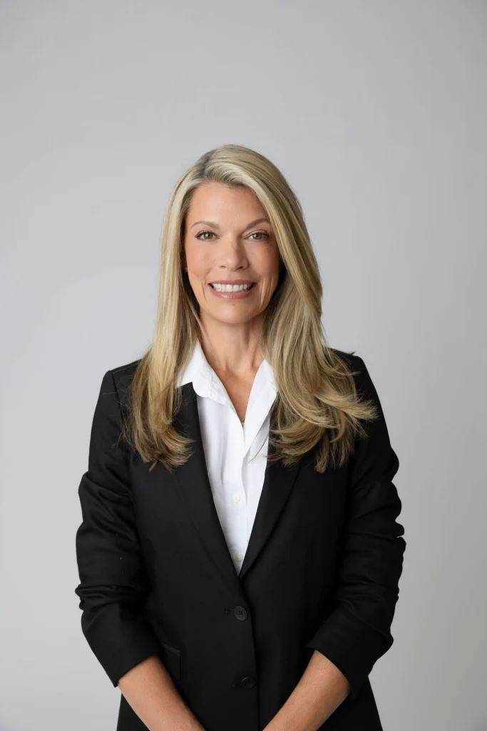 Professional woman with blonde hair in a black blazer and white blouse smiling against a neutral gray background